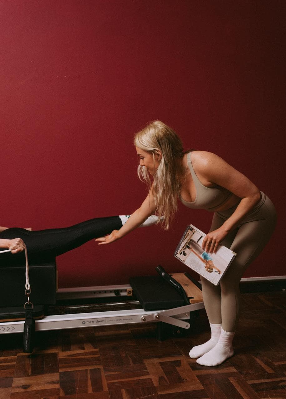 An instructor leans over a reformer to correct a client's extended leg, a manual held open beside her.