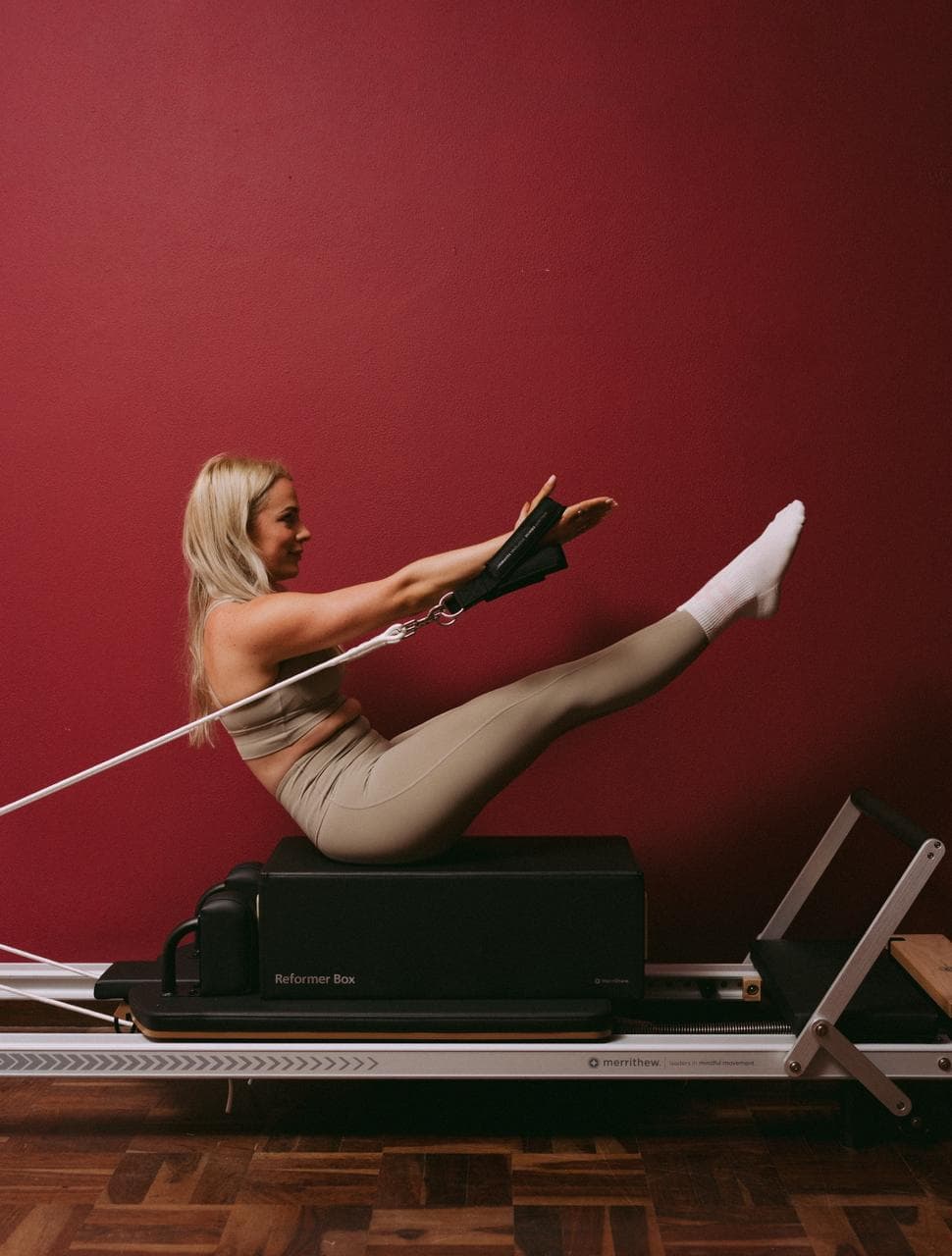 A teaser held on the reformer box, legs extended and arms reaching along the straps.
